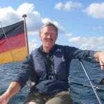 Foto von Dieter Peters-Kühnel, einem Mann mit heller Haut, kurzen weißen Haaren und einem braunen Schnurrbart. Er trägt einen braunen Pullover und reine blaue Schwimmweste und blickt lächelnd in die Kamera, während er an einer Reling von einem Boot sitzt.