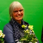 Foto von Heike Arndt, einer Frau mit heller Haut und blonden Haaren. Sie trägt eine runde schwarze Brille, ein blaues Shirt und einen blauen Schal mit weißen Punkten und blickt lächelnd in die Kamera, die Hände halten einen Blumentopf mit hellen Blumen.