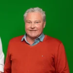 Foto von Kurt Schäfer, ein Mann mit heller Haut und kurzen grauen Haaren. Er trägt ein blaukariertes Hemd und einen roten Strickpullover und blickt lächelnd in die Kamera.