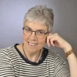 Foto von Susanne Ibold, einer Frau mit heller Haut und kurzen grauen Haaren. Sie trägt eine dunkle runde Brille, eine weißen Strickpullover mit schwarzen Streifen und blickt lächelnd in die Kamera, eine Hand stützt locker den Kopf.